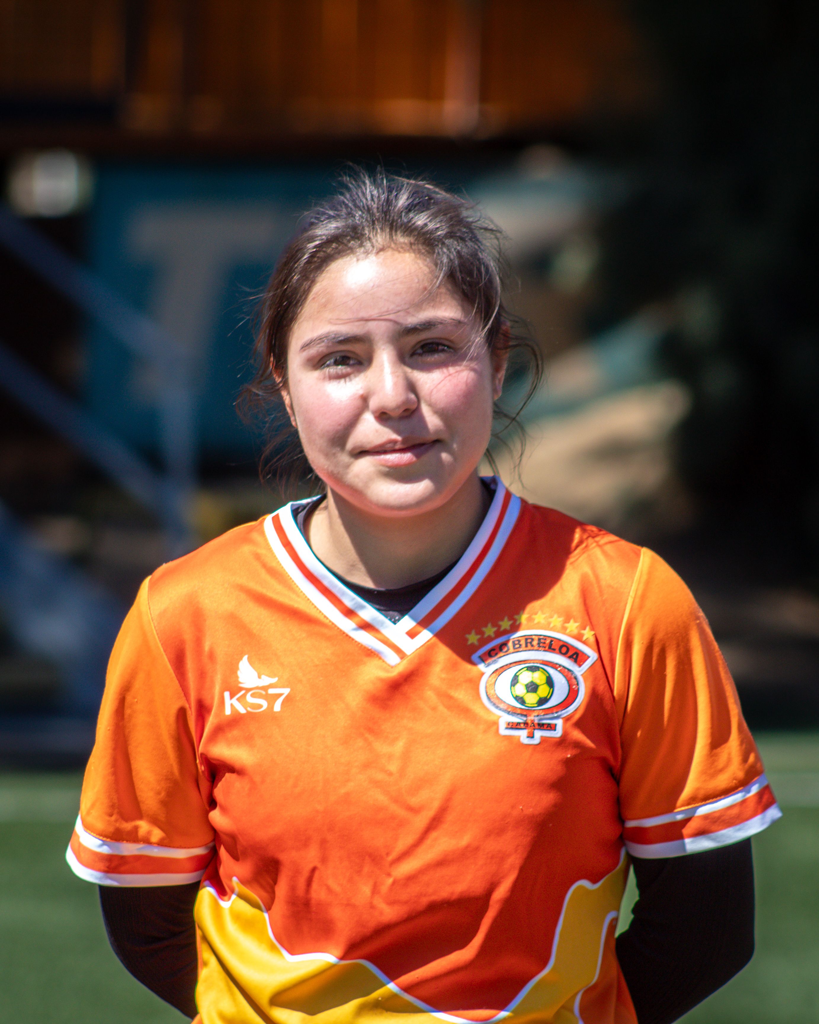 Portrait of a young female footballer in an orange jersey standing on a sunny field, exuding confidence and determination.