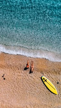 Aerial view of two adults relaxing on a sandy beach with a yellow surfboard by clear blue waters.