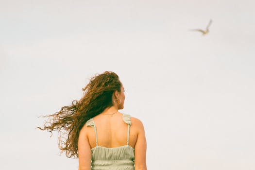 A woman stands peacefully with her hair in the wind, gazing at the sky, evoking tranquility.