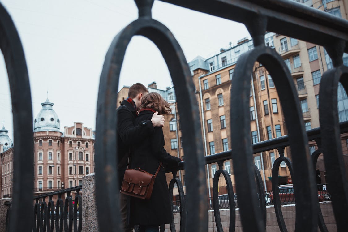 From below of anonymous female and male in warm clothes standing near railing together and hugging in cold weather