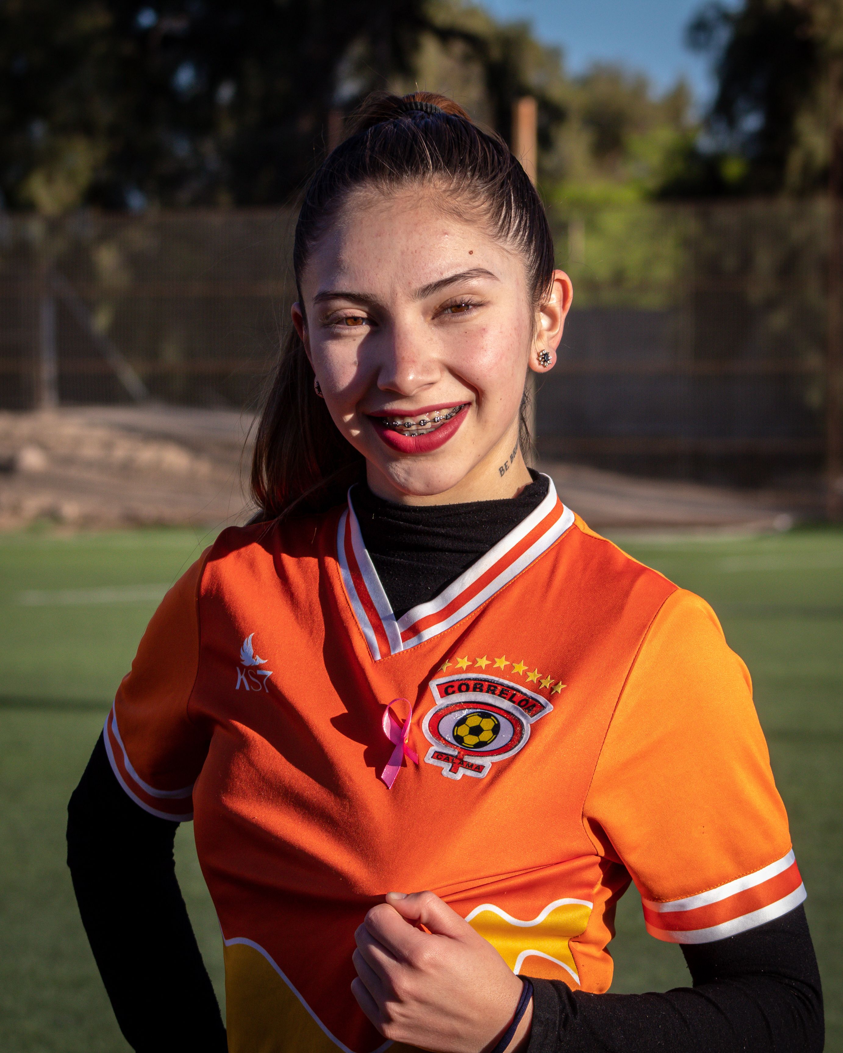 A young woman wearing an orange soccer jersey smiles confidently on a sunny day at the football field.