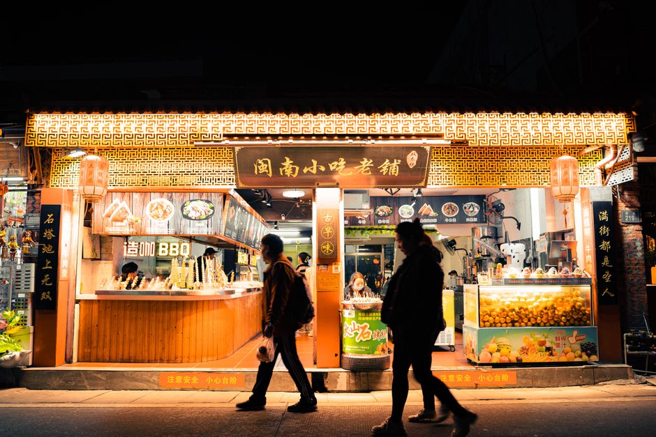Lively street food stall in a bustling night market with people passing by and bright neon signs.