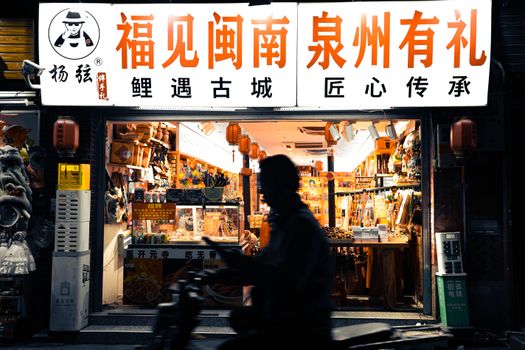 Silhouette of a person passing a vibrant night market shop in Quanzhou, China.