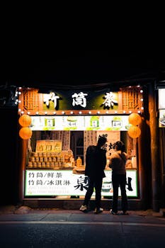 Two people explore a vibrant tea shop in Asia, illuminated by lanterns at night.