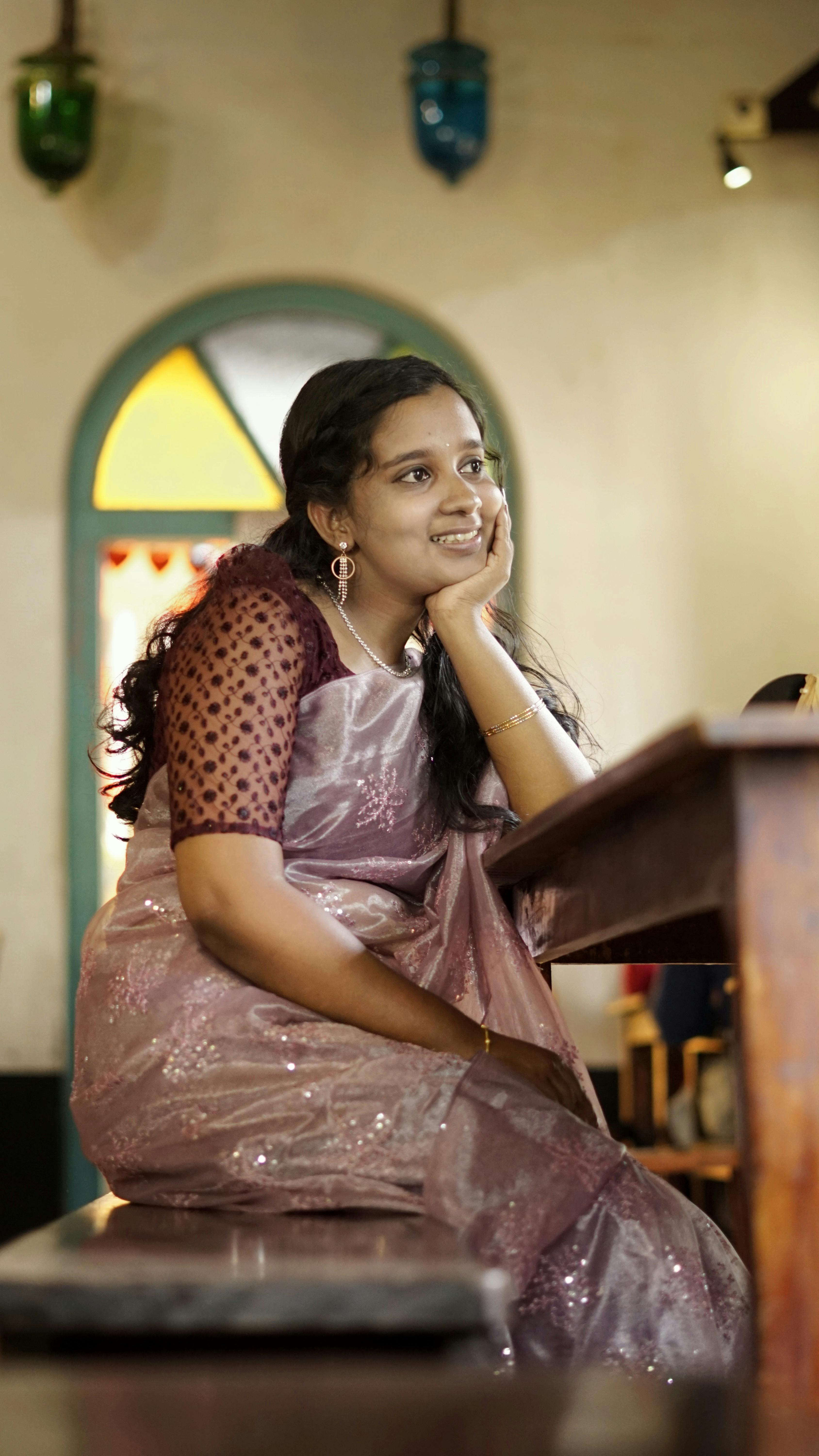 Young woman in a stylish saree sitting indoors, gazing thoughtfully.