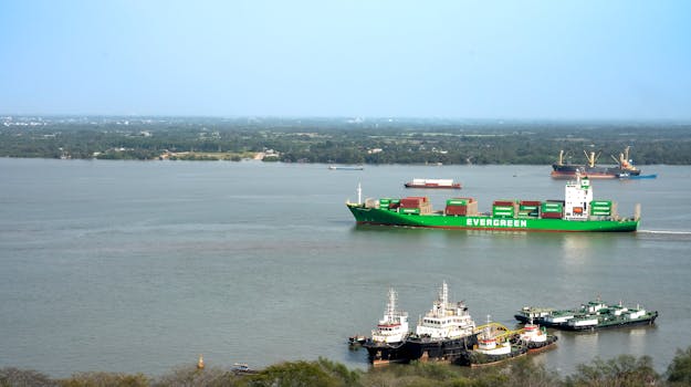 Cargo ships navigate a river with a lush landscape in the background.