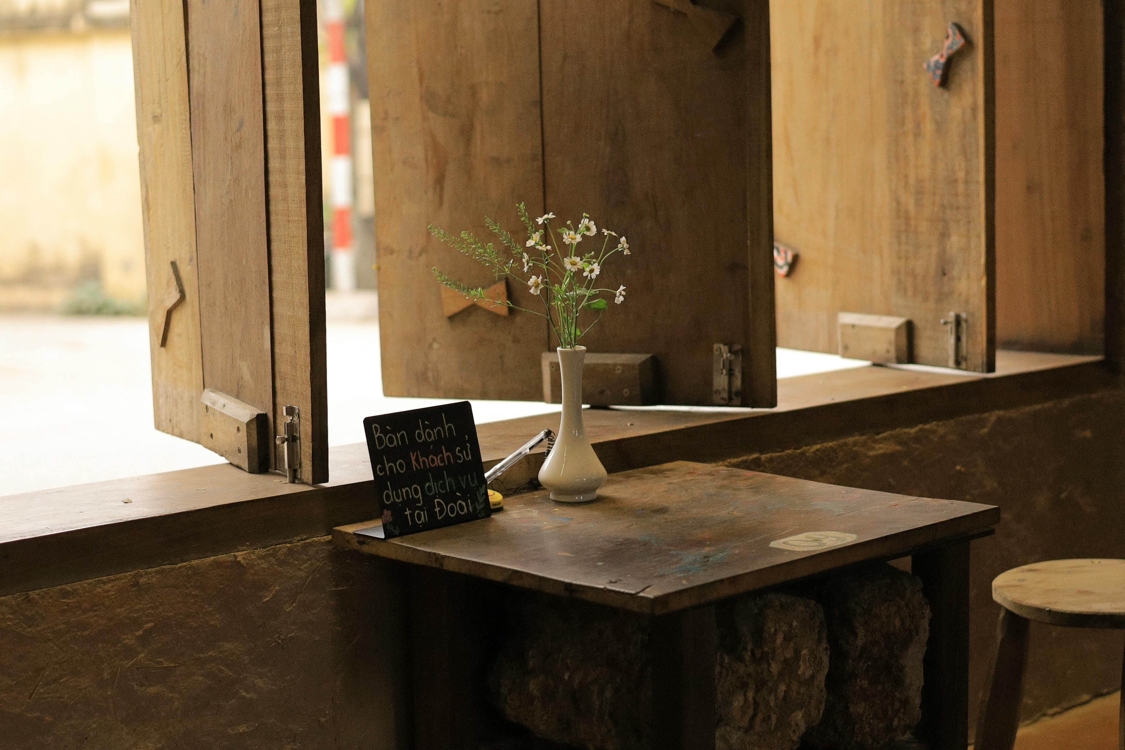 Gratuit O atmosferă rustică și intimă de cafenea, cu o masă de lemn, flori într-o vază și un semn decorativ într-o fereastră însorită. Fotografie de stoc