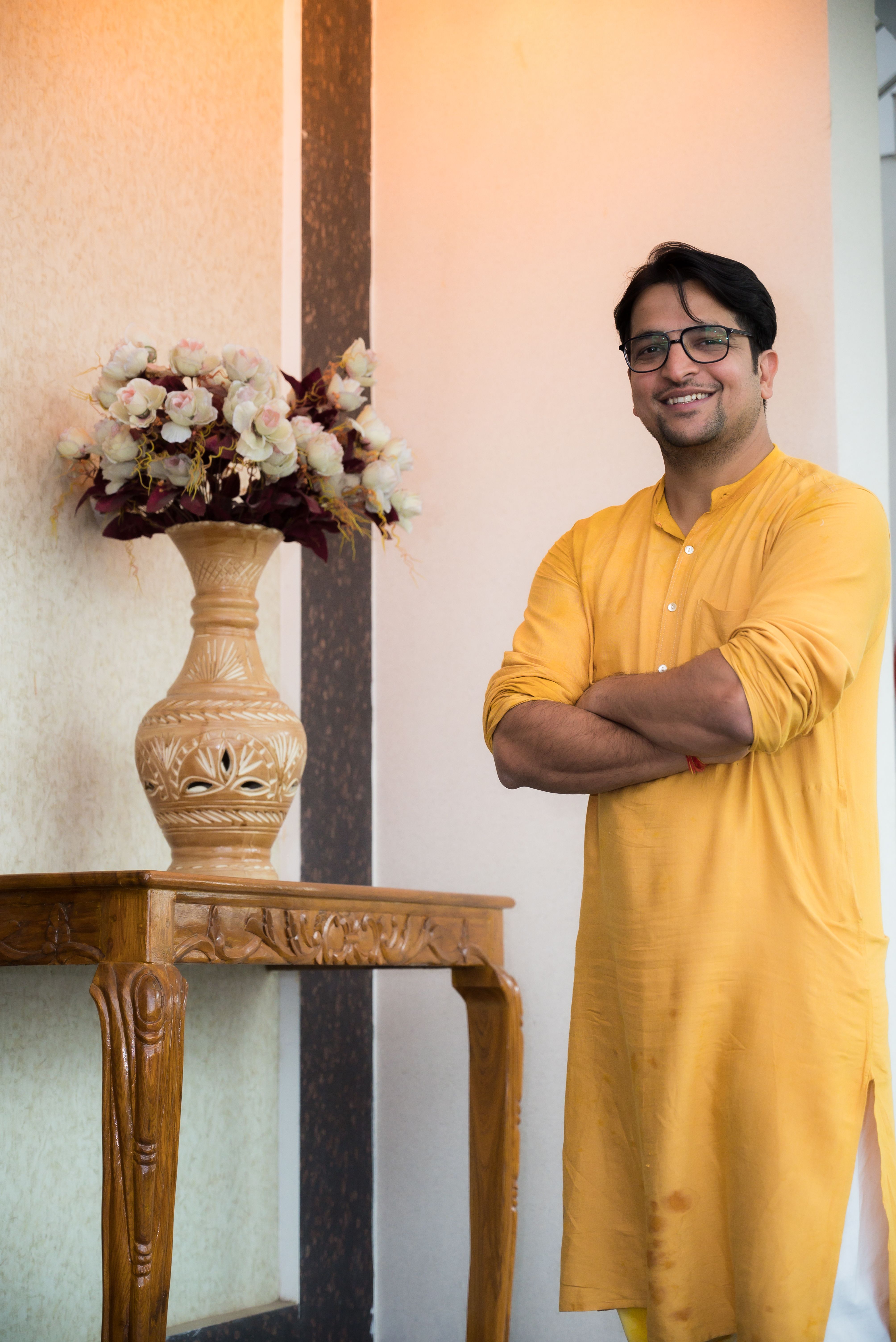 Smiling man in a yellow kurta stands beside a decorative vase on a wooden table indoors.