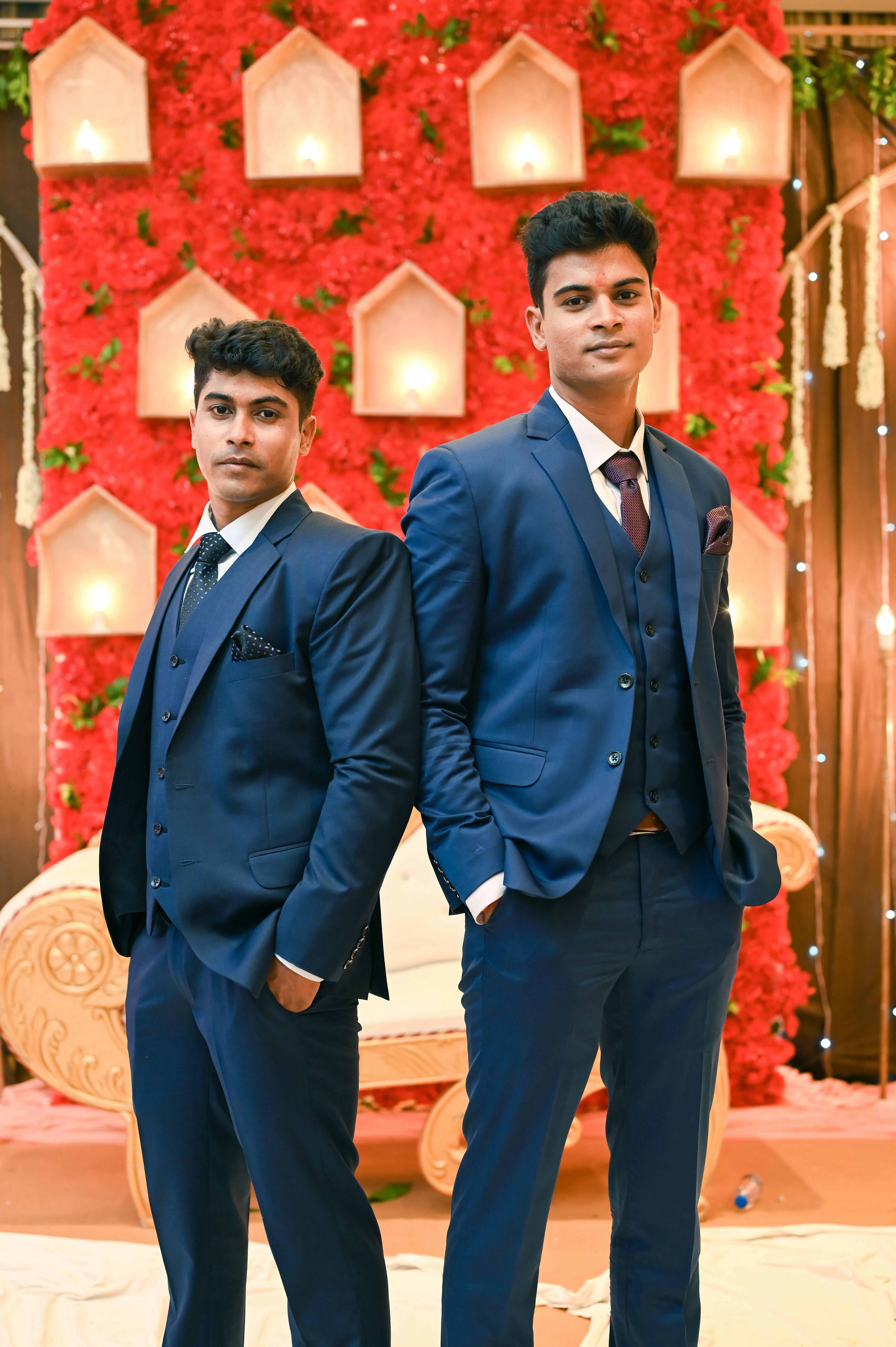 Two groomsmen in blue suits pose elegantly in front of a lavish wedding backdrop.