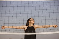 一位身着黑色连衣裙的时尚女子，在晴朗的蓝天下，于排球网后优雅地摆出姿势