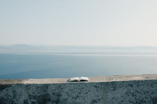 A solitary open book rests on a ledge with a serene sea view and clear skies.