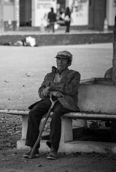 Black and white portrait of a senior man resting on a bench outdoors with a cane.
