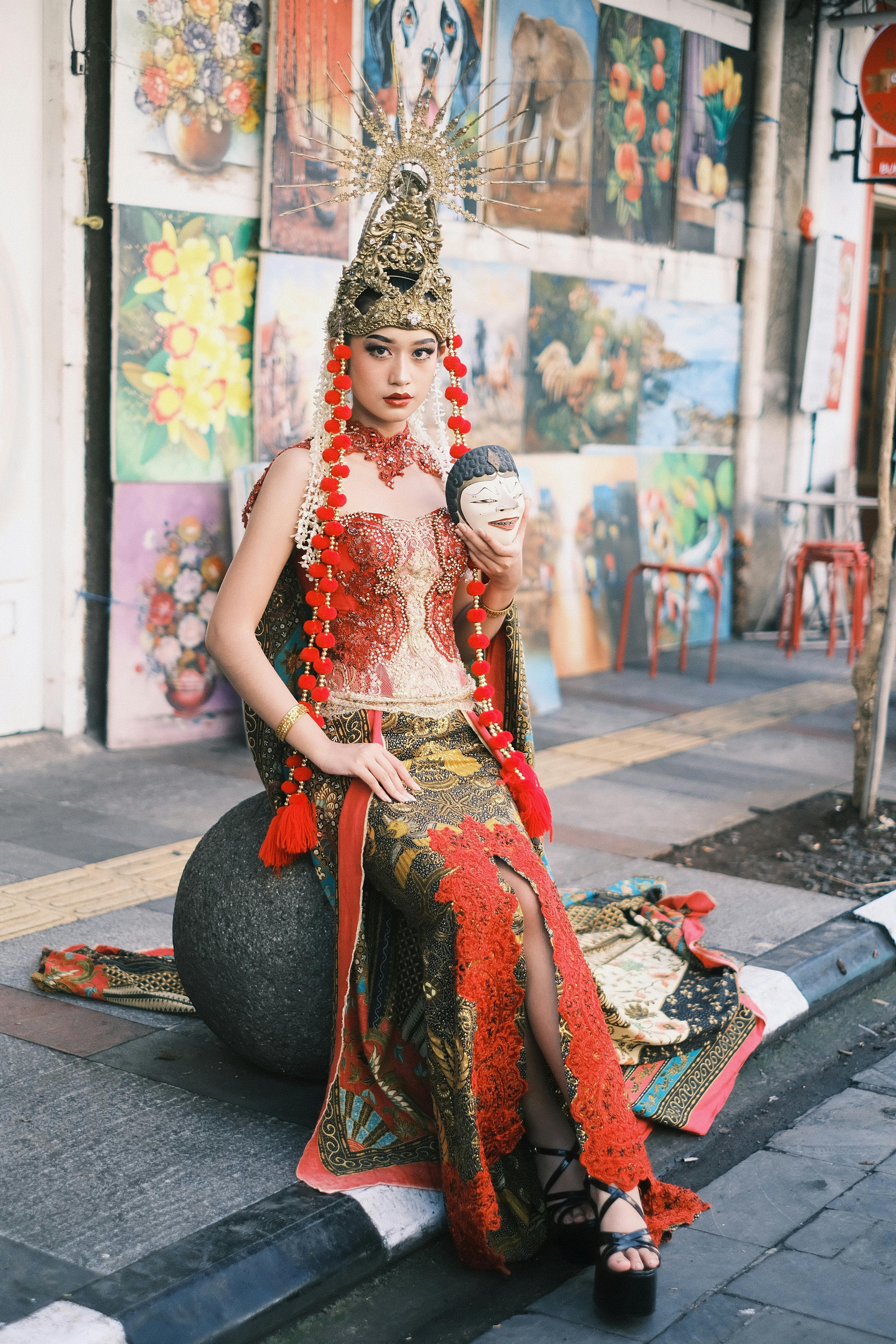 Woman in traditional Indonesian attire with mask, sitting in vibrant street setting. Captivating cultural display.