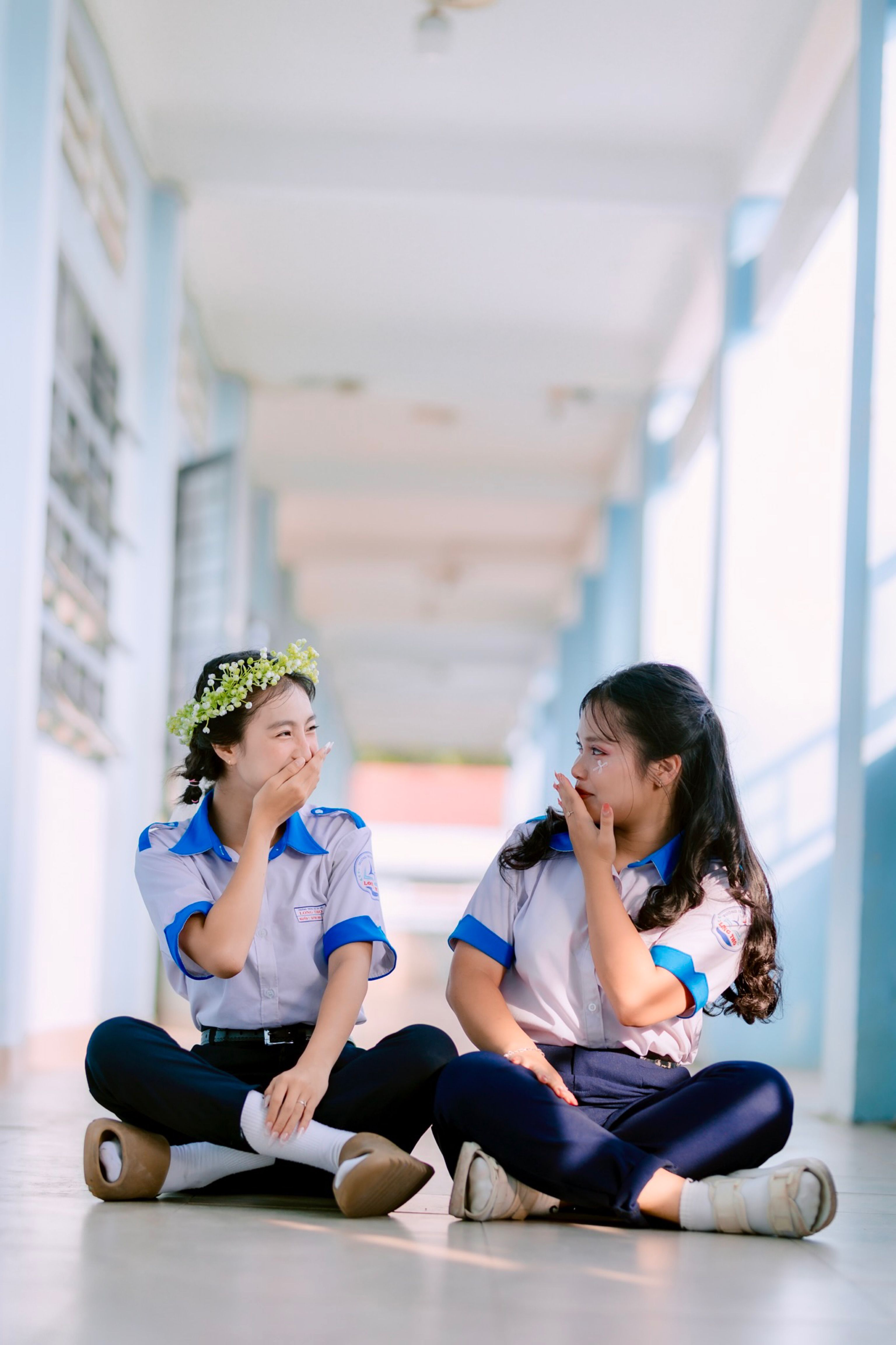 Two schoolgirls sit laughing in a sunlit school hallway, enjoying a carefree moment.