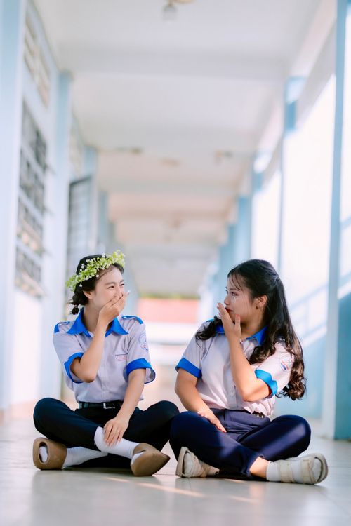 Free Two schoolgirls sit laughing in a sunlit school hallway, enjoying a carefree moment. Stock Photo