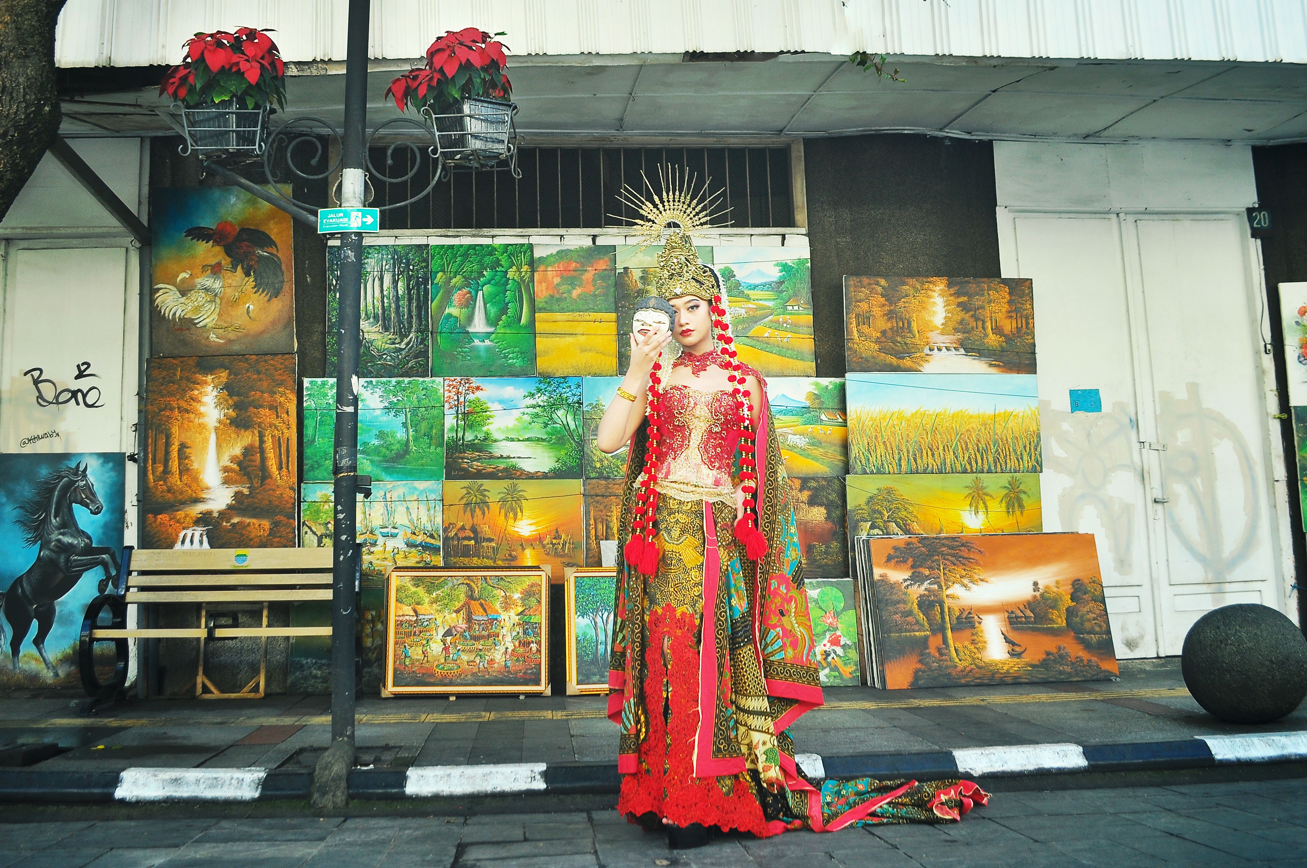 A Balinese dancer in traditional attire poses in front of vibrant paintings on a street.