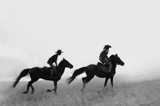 Two riders on horseback galloping across a misty plains, captured in dynamic black and white.