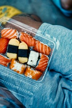 Close-up of assorted sushi, including salmon, tamago, and maki, in a plastic container on denim shorts.