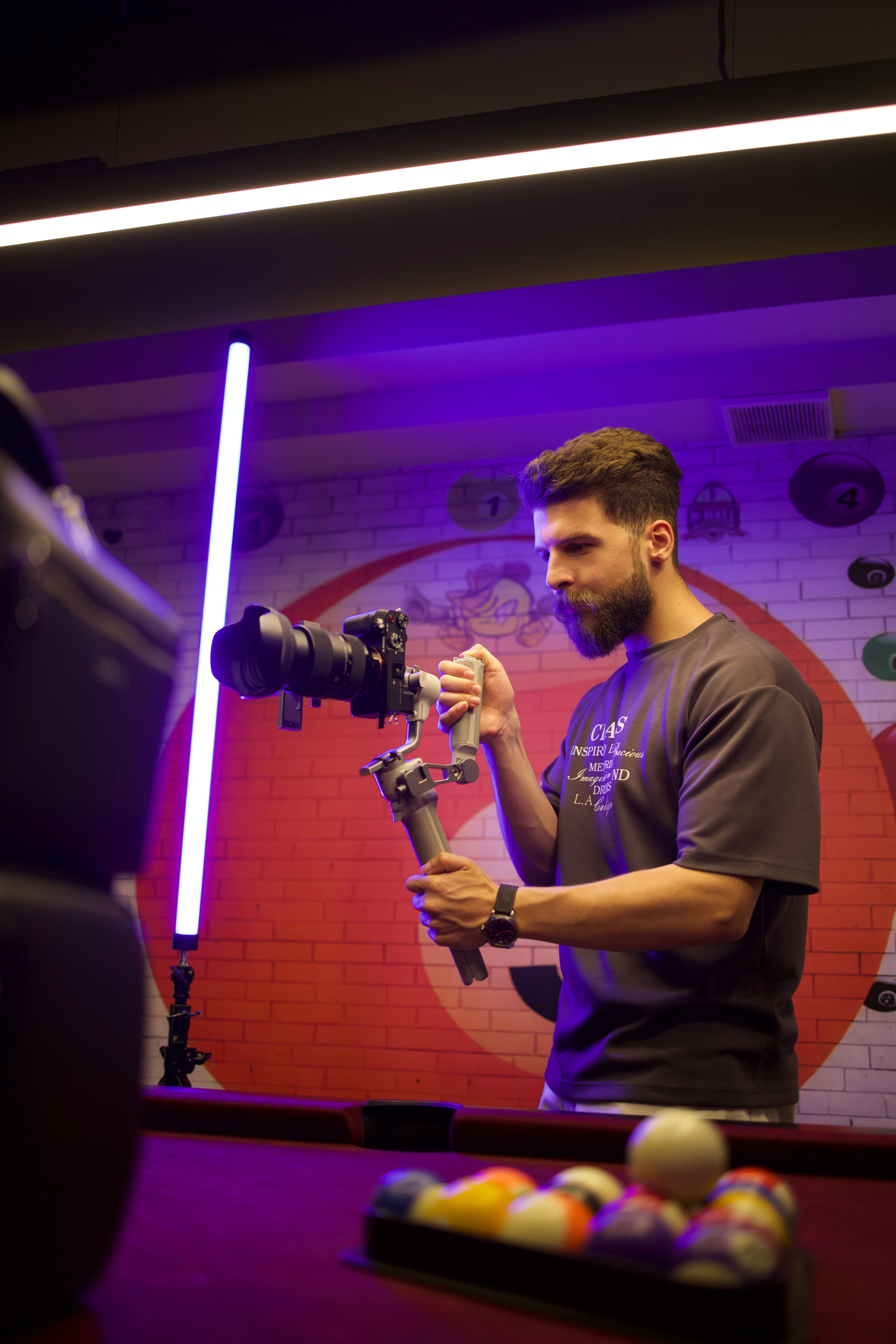Man using a camera stabilizer for filming indoors with neon lights and billiard table.