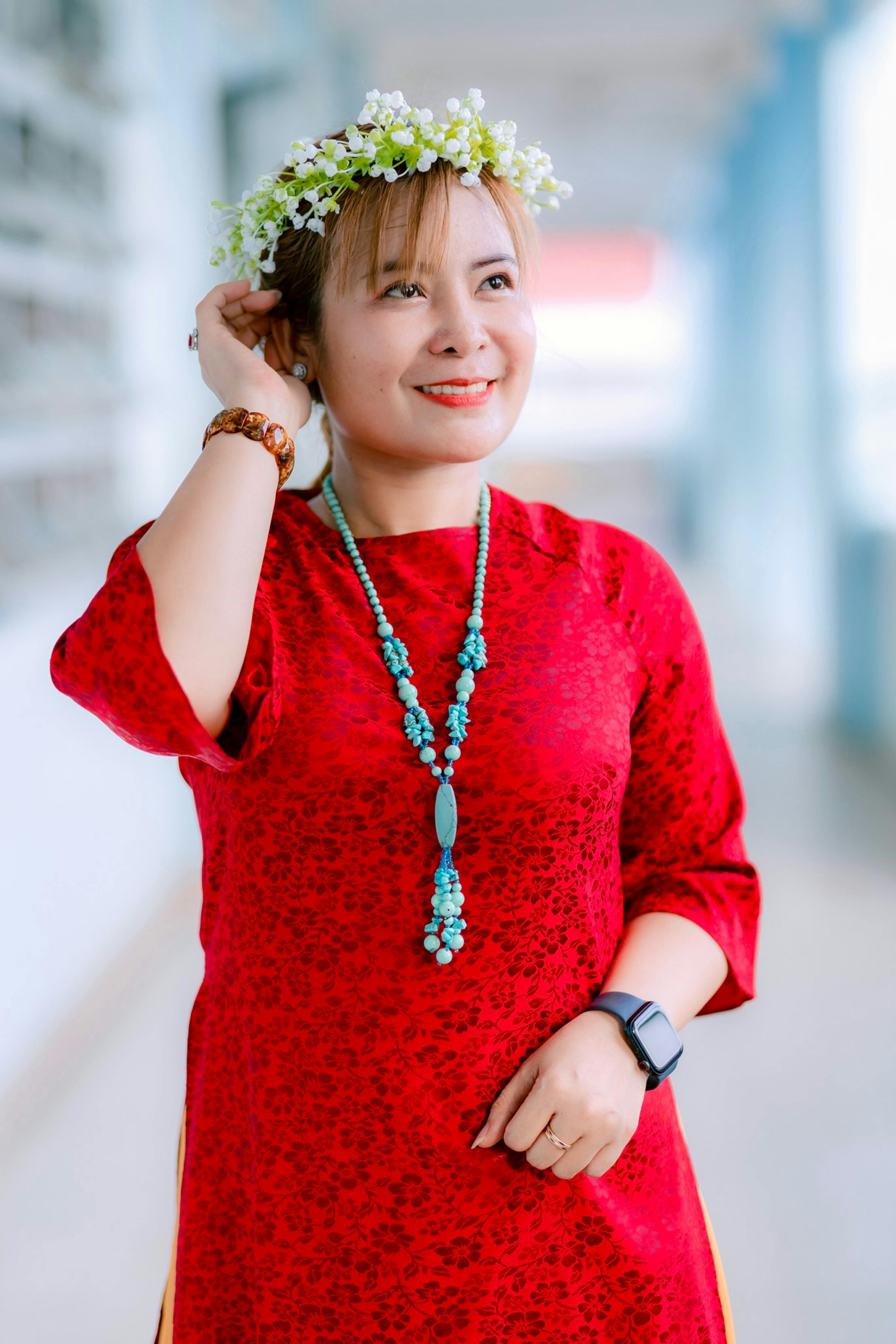 Woman in a red dress wearing a floral crown and turquoise necklace smiles outdoors.