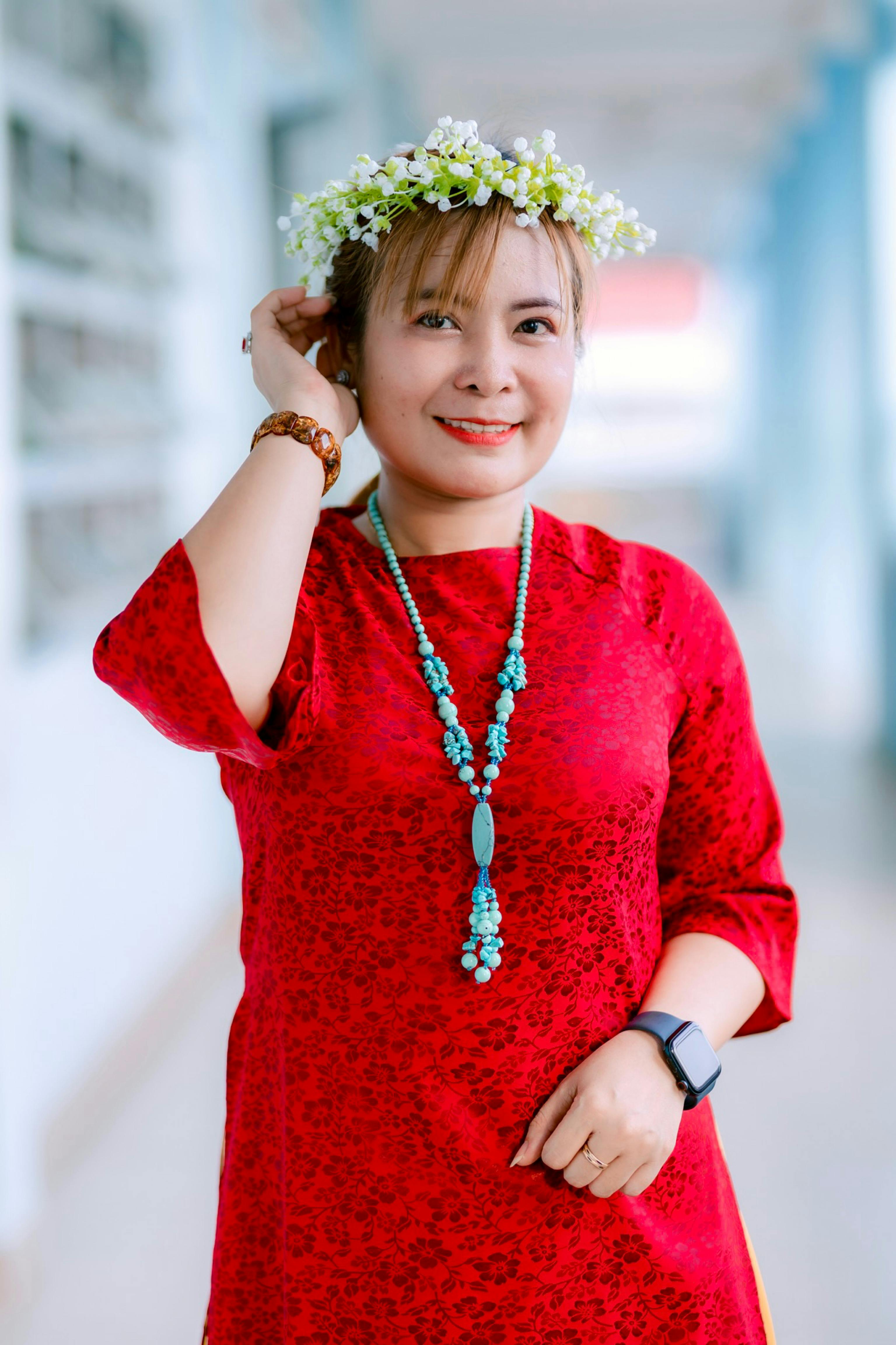 Smiling woman in floral crown and red dress adjusting a necklace outdoors.