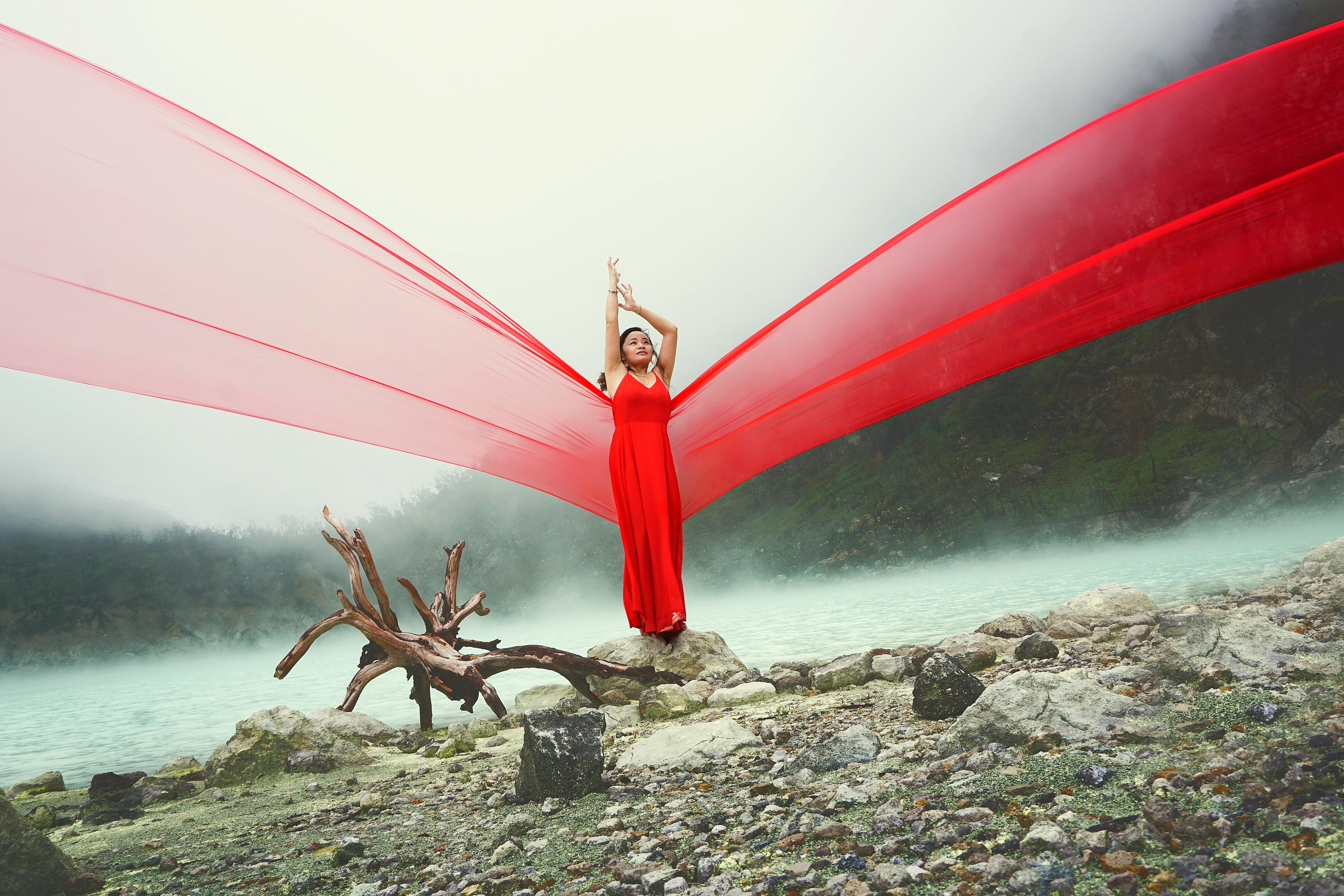 A woman in a red dress poses by a misty turquoise lake with dramatic fabric flow.