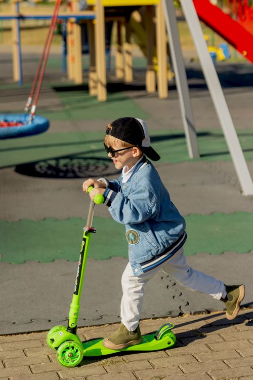 Free A playful child rides a green scooter outdoors on a sunny day, wearing a denim jacket and cap. Stock Photo
