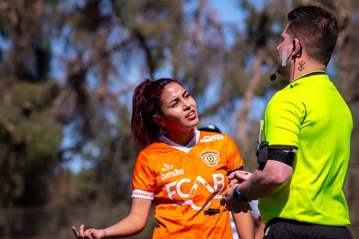 Female soccer player in orange jersey argues with referee on the outdoor field during a sunny day match.