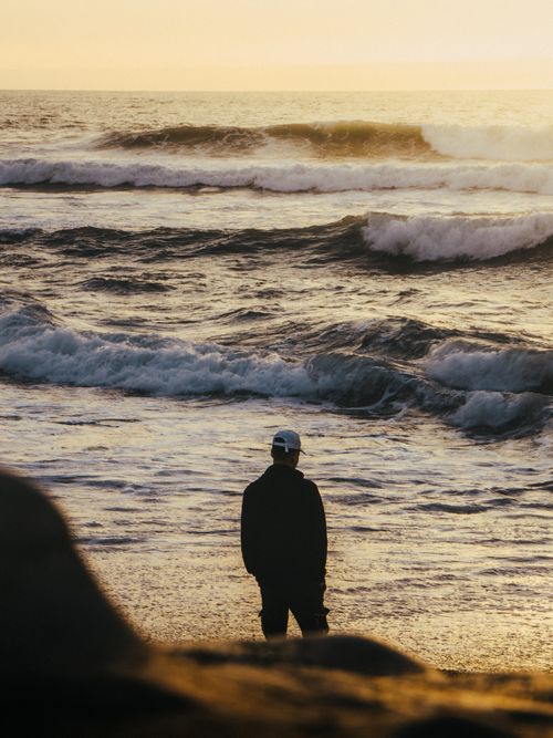 Free A lone person gazes at waves on a Moroccan beach during sunrise, evoking tranquility. Stock Photo