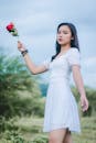 一位身穿白色长裙的年轻女子，手持红玫瑰，神情安详地站在户外