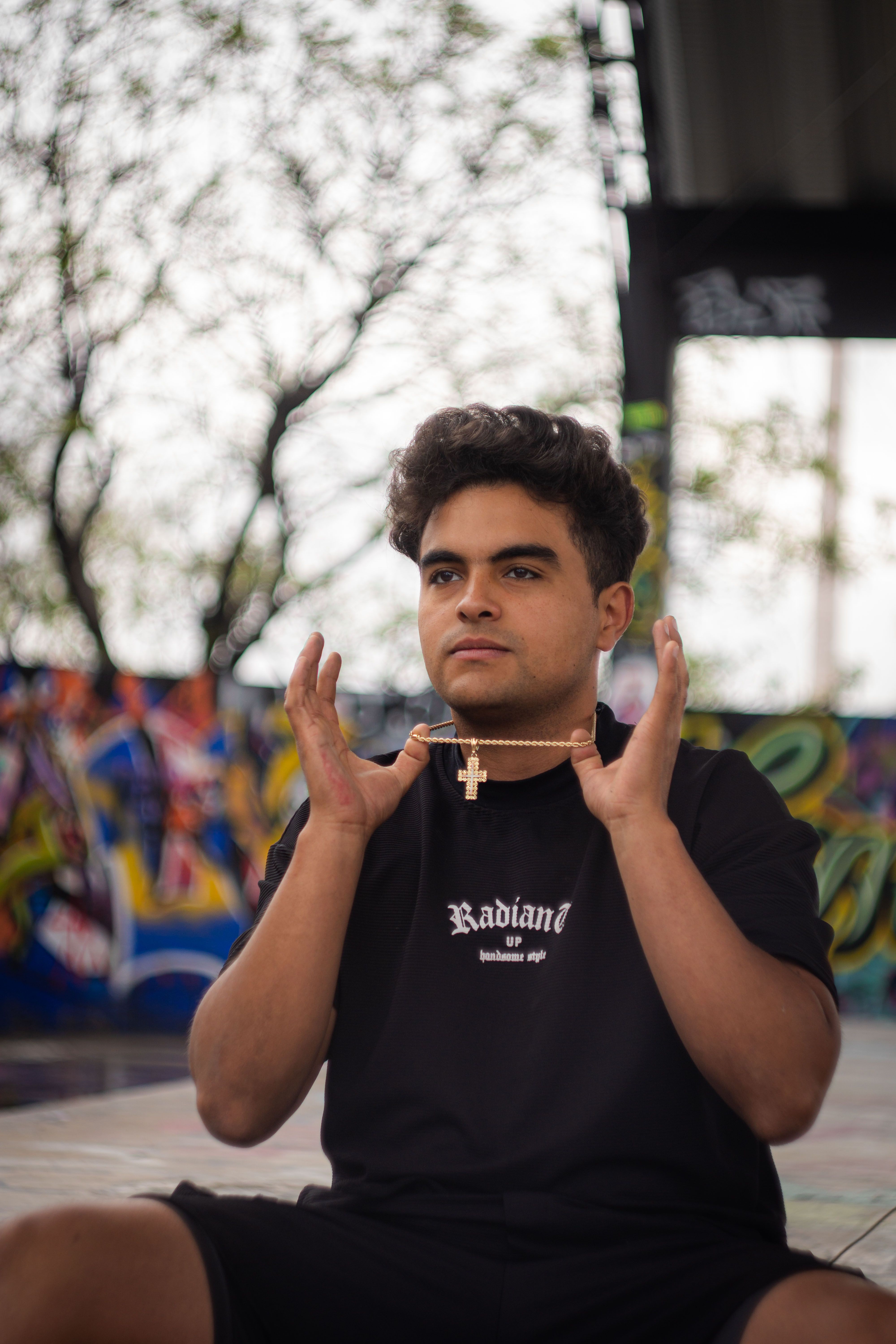 A young man holding a gold cross necklace in an outdoor urban setting with vibrant graffiti art.