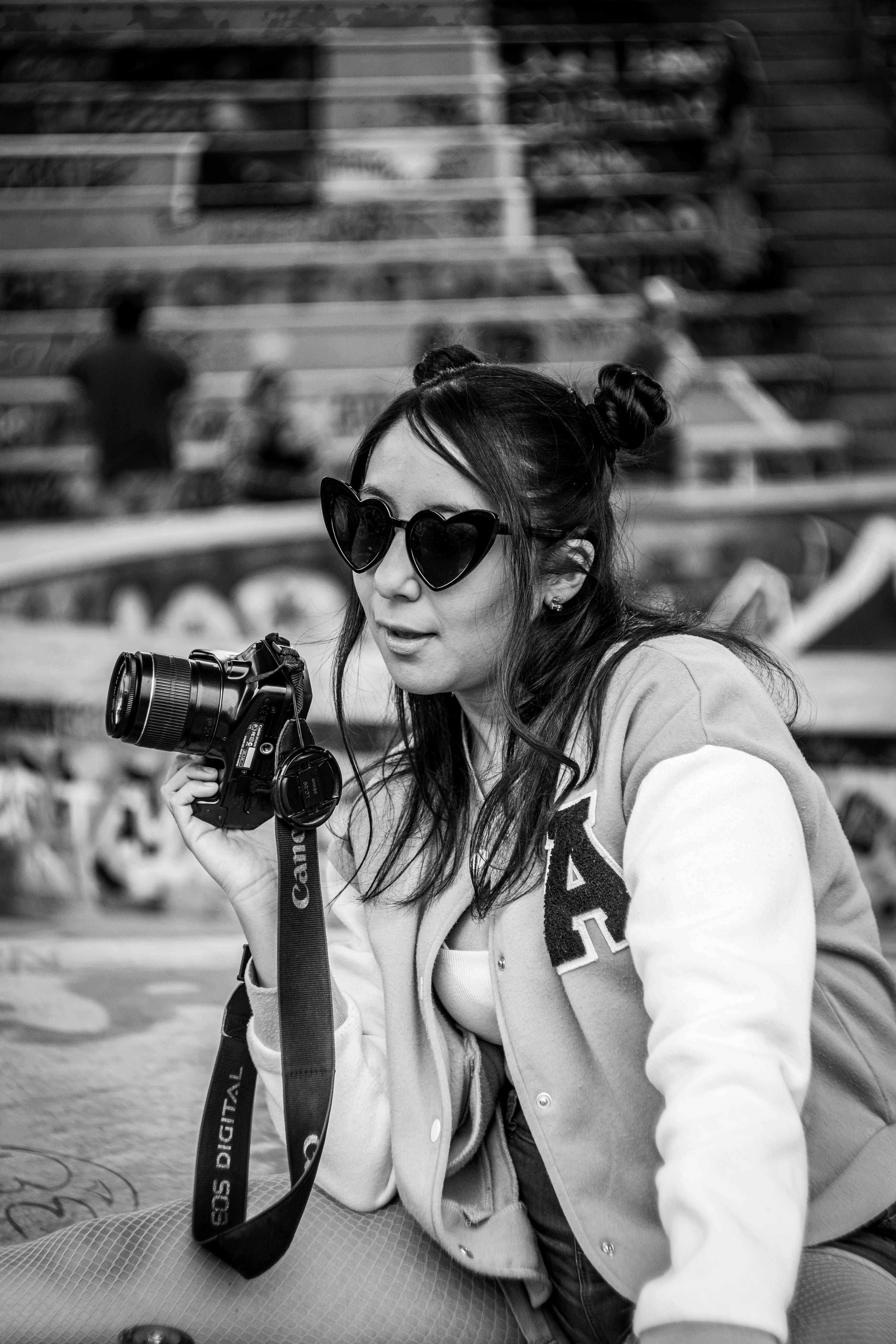 Black and white portrait of a stylish woman photographer with sunglasses at an urban skate park, holding a camera.