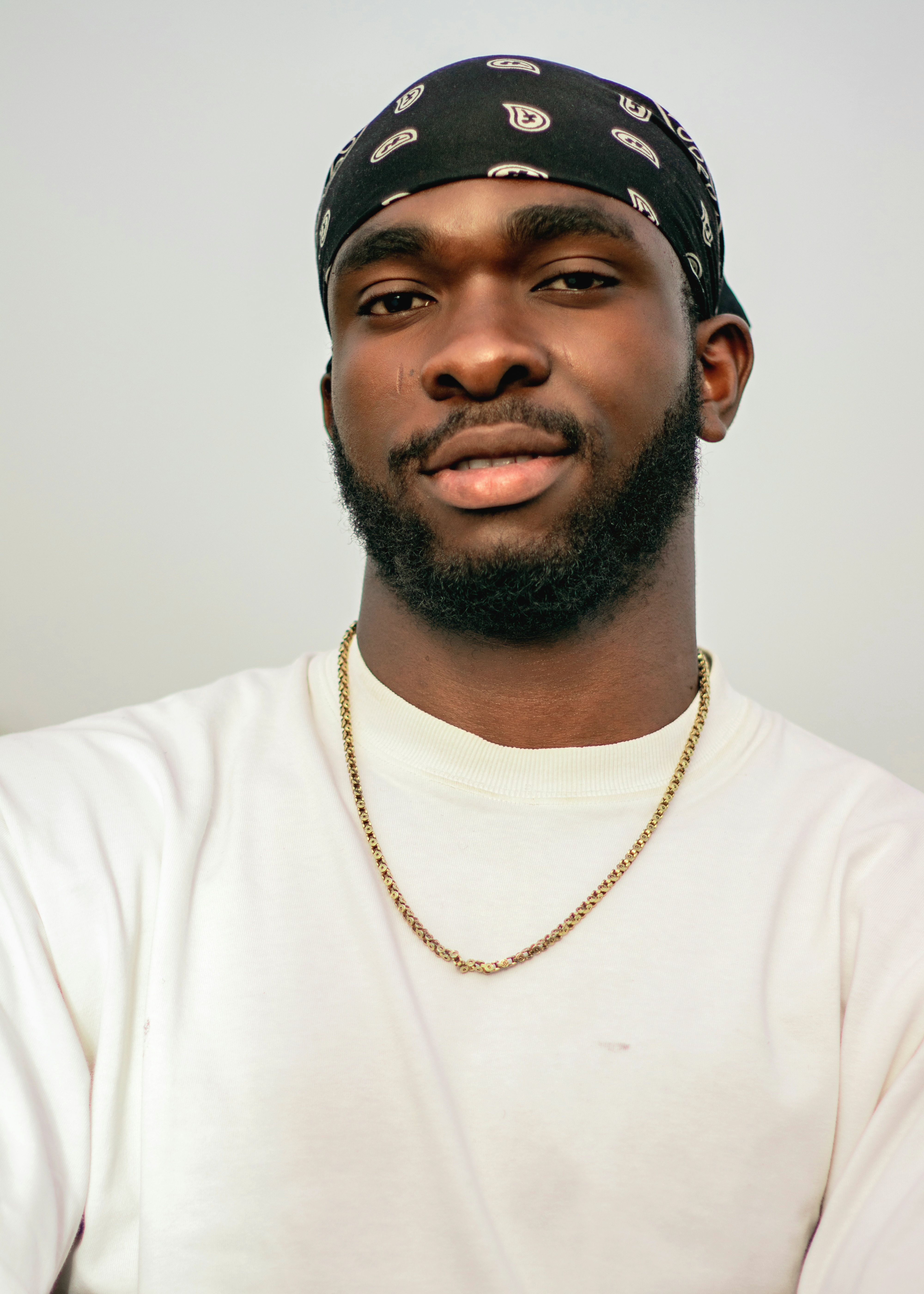 A young man wearing a black bandana and chain necklace, exuding confidence and style in a simple indoor setting.