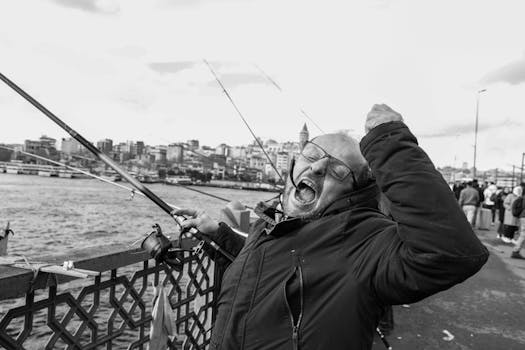 Excited fisherman celebrates catch on Istanbul's Galata Bridge, with cityscape background, in black and white.