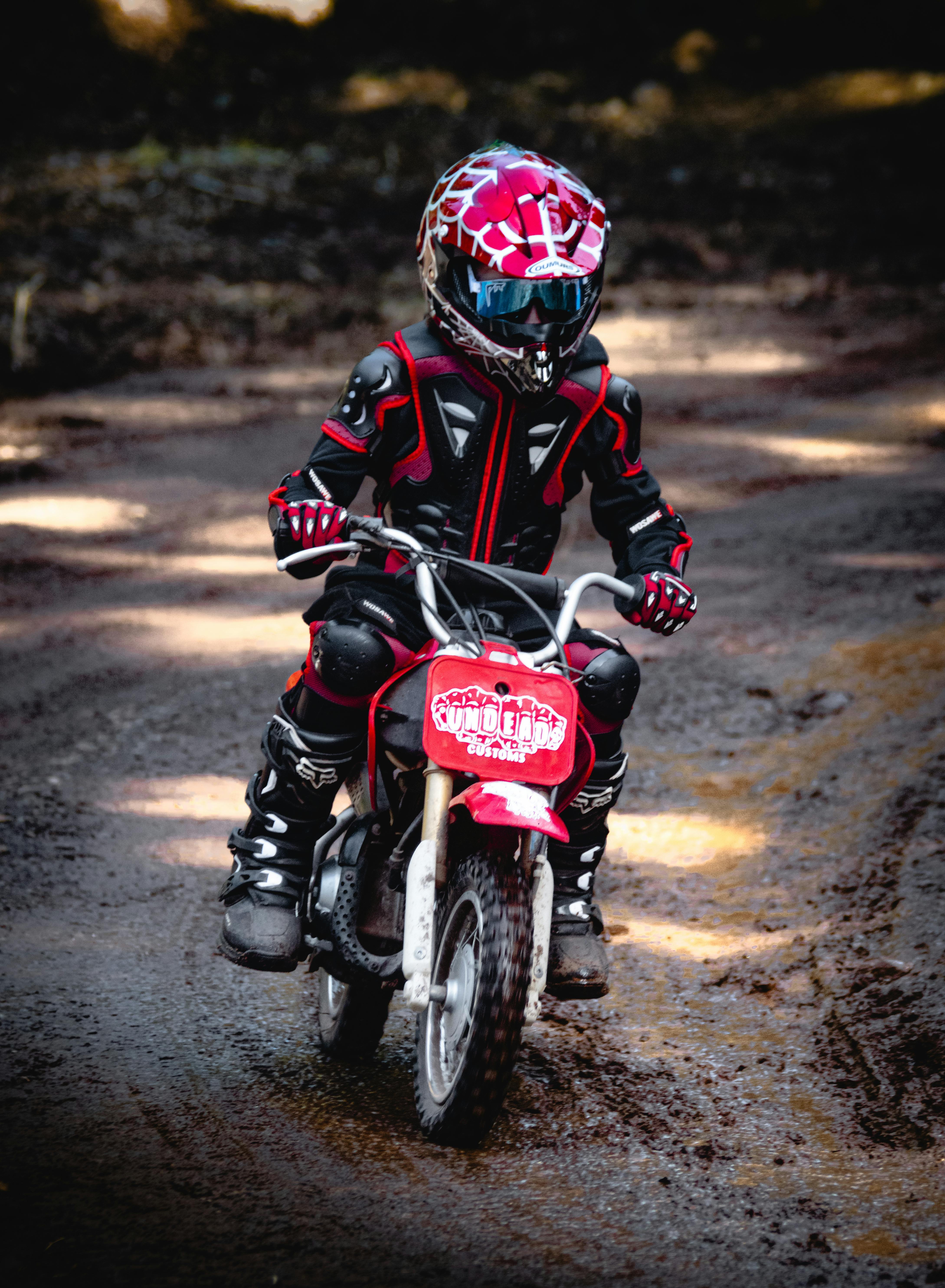 Young child in protective gear riding a mini dirt bike on a wooded trail.