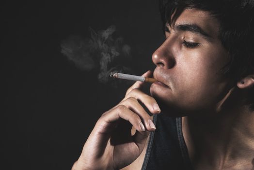 Close-up of a young man smoking a cigarette against a dark background, emphasizing smoke.