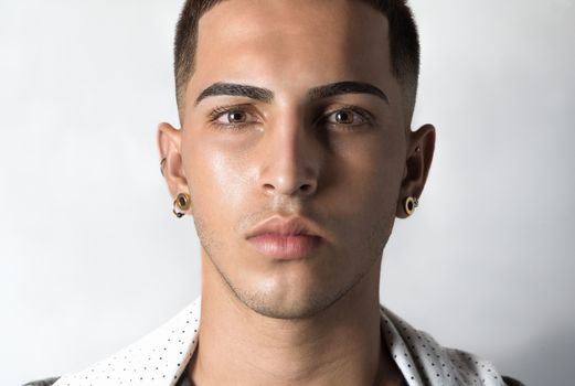 Close-up portrait of a young man with earrings and sharp features, looking directly at the camera.