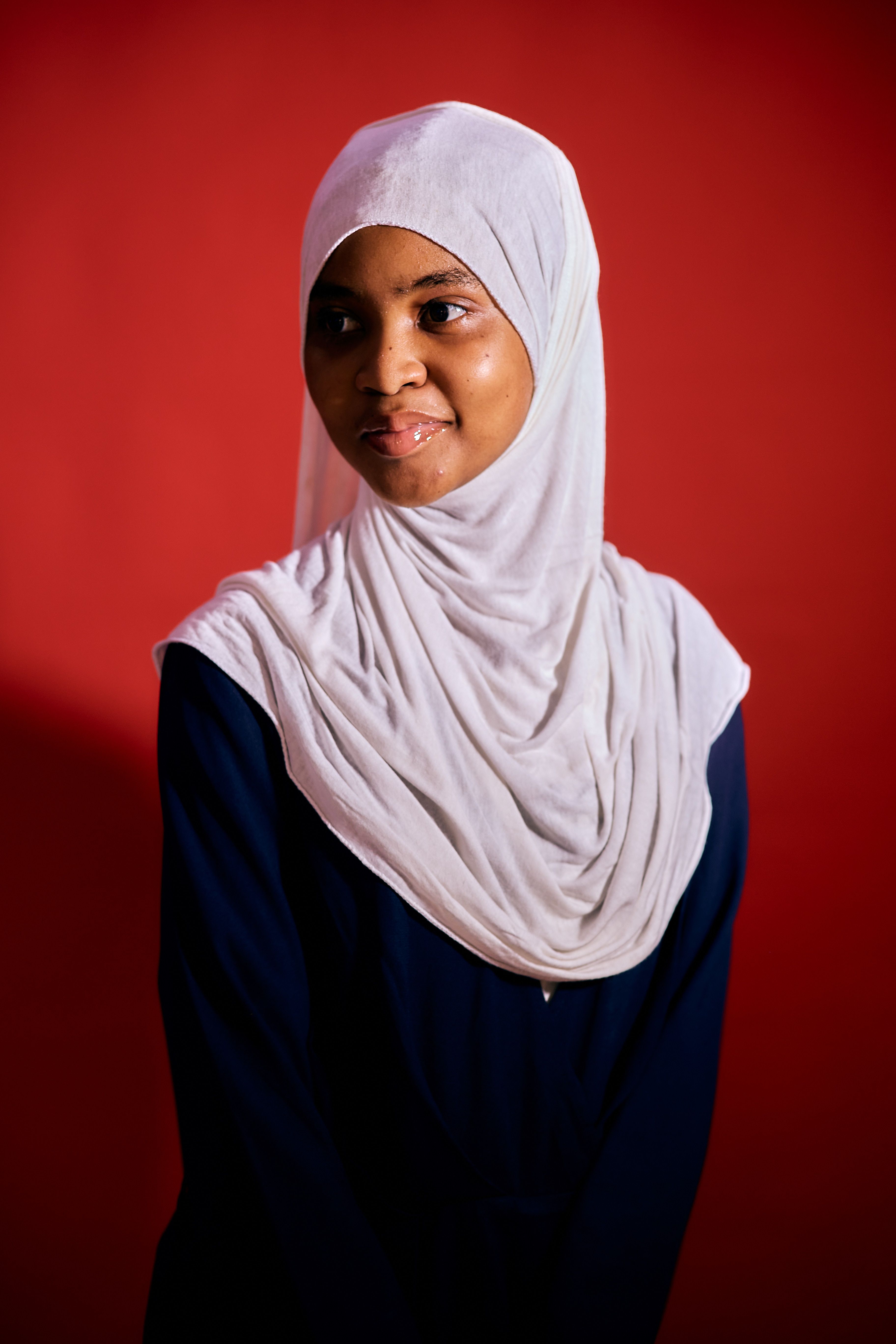 A thoughtful portrait of a woman wearing a white hijab, set against a vibrant red background.