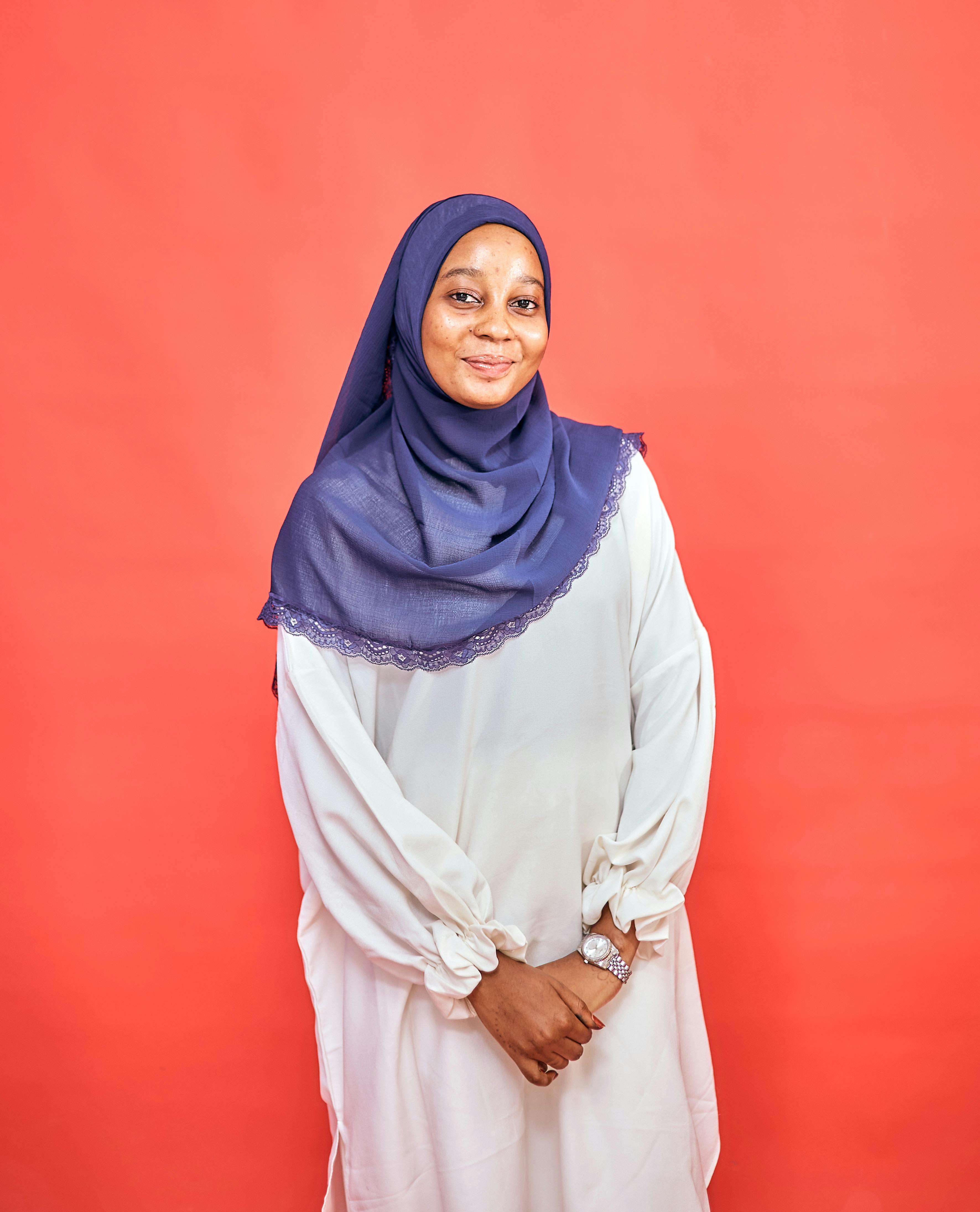 Smiling woman wearing a blue hijab and white dress against a vibrant red backdrop