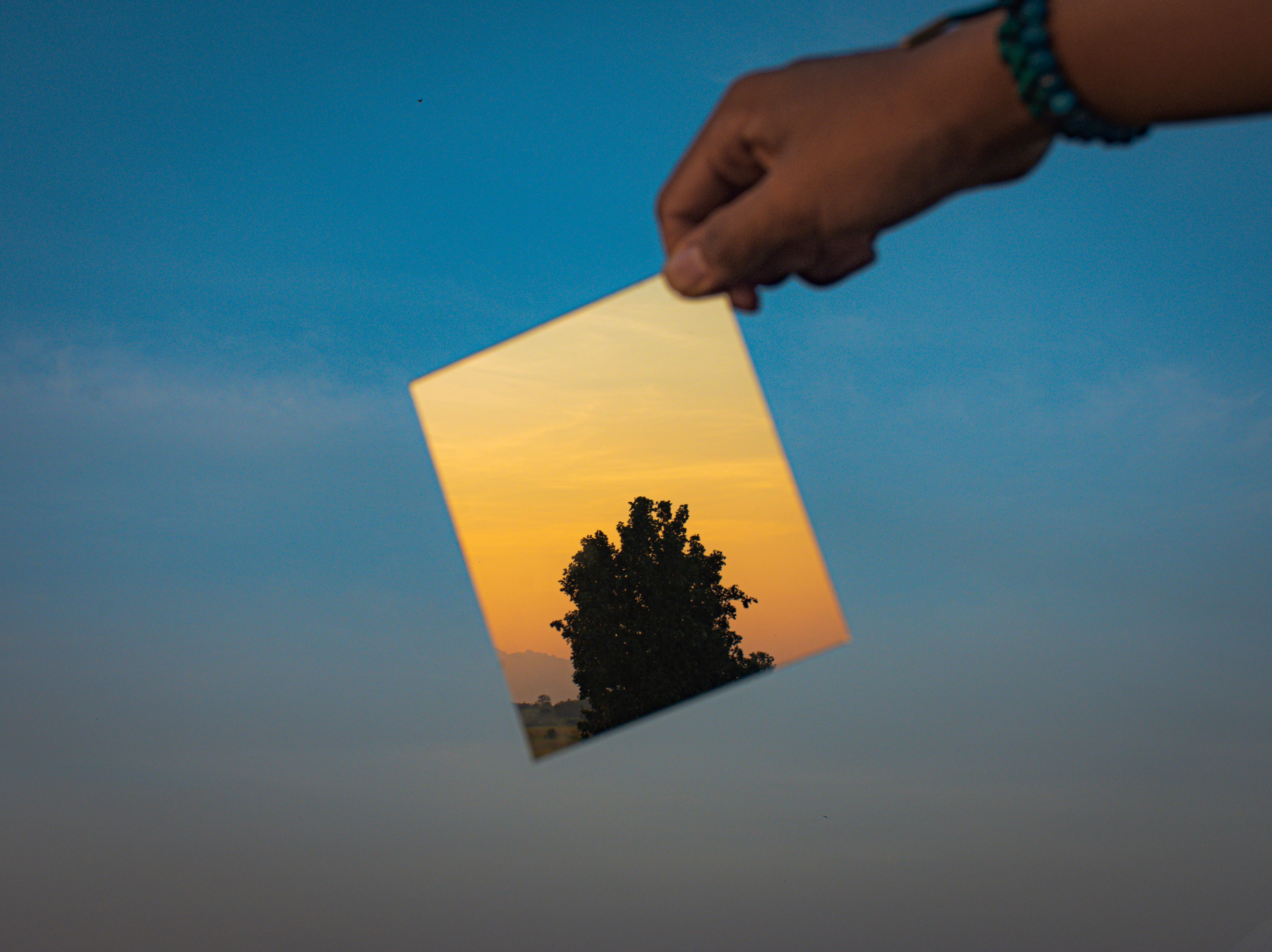 Hand holding a mirror reflecting a tree against a sunset sky in Thailand.