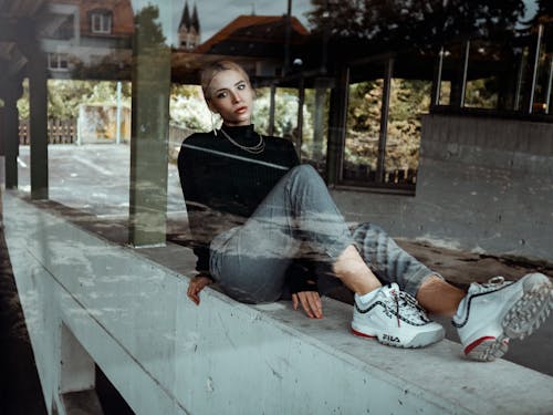 Through glass of serious stylish young female in trendy outfit sitting on concrete underground block and looking at camera