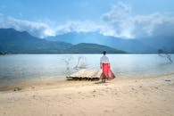 一名女子身着红裙，站在宁静的海滩上，凝视着大海，背景是连绵的山脉