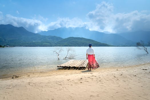 一名女子身着红裙，站在宁静的海滩上，凝视着大海，背景是连绵的山脉
