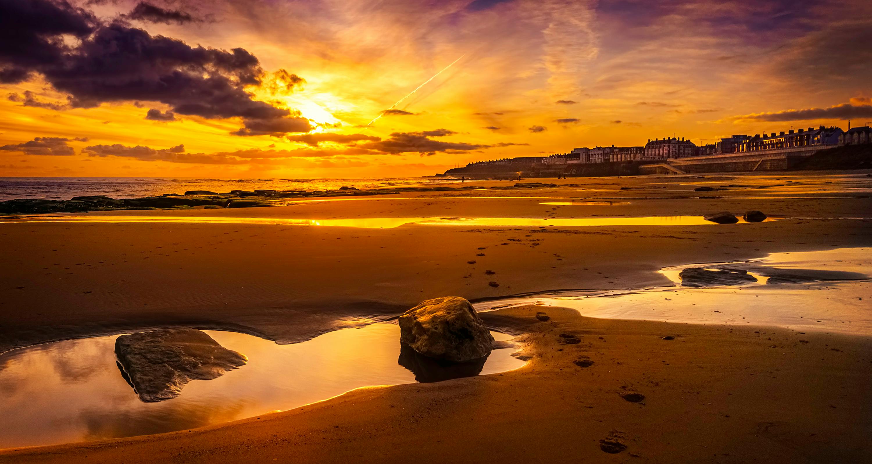 宁静的海滩上，金色的日落映在沙滩倒影中，远处是海岸线的建筑