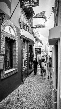 Black and white street scene in Santorini with people walking under tavern signs.