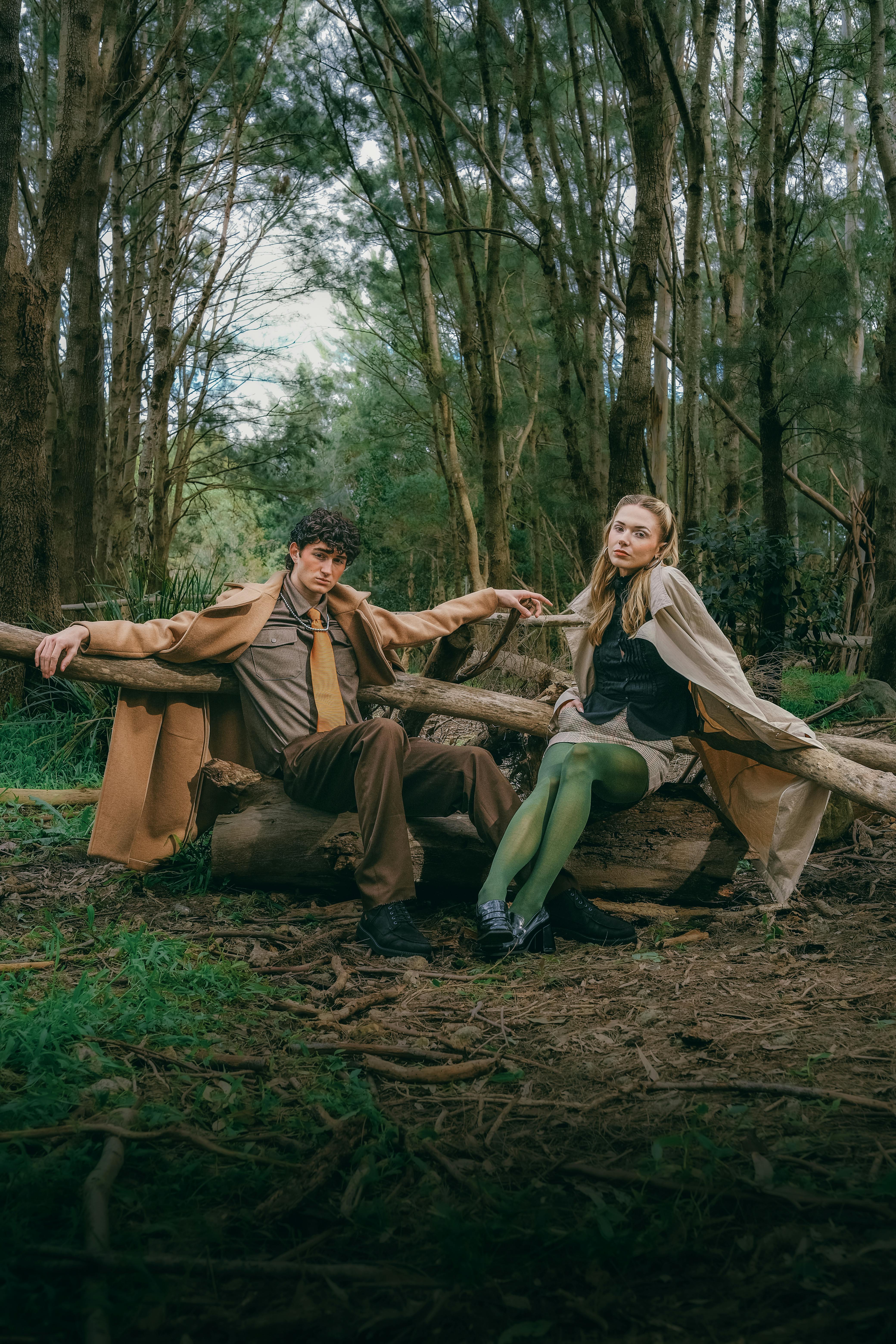A fashionable couple sitting on logs amidst a dense green forest, exuding a vintage vibe.