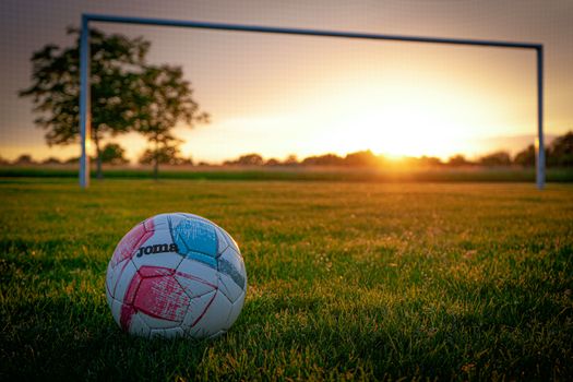 A soccer ball on a grassy field with a goal post, beautifully lit by the vibrant sunset.