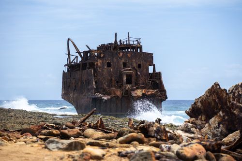 Free A rusting shipwreck on Curaçao's rocky coast with waves crashing, under a clear blue sky. Stock Photo