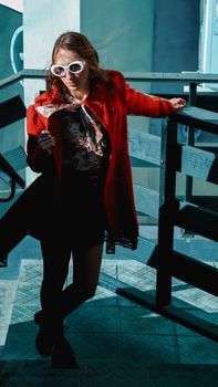 Fashionable woman in red coat with sunglasses posing on urban stairs in sunlight.