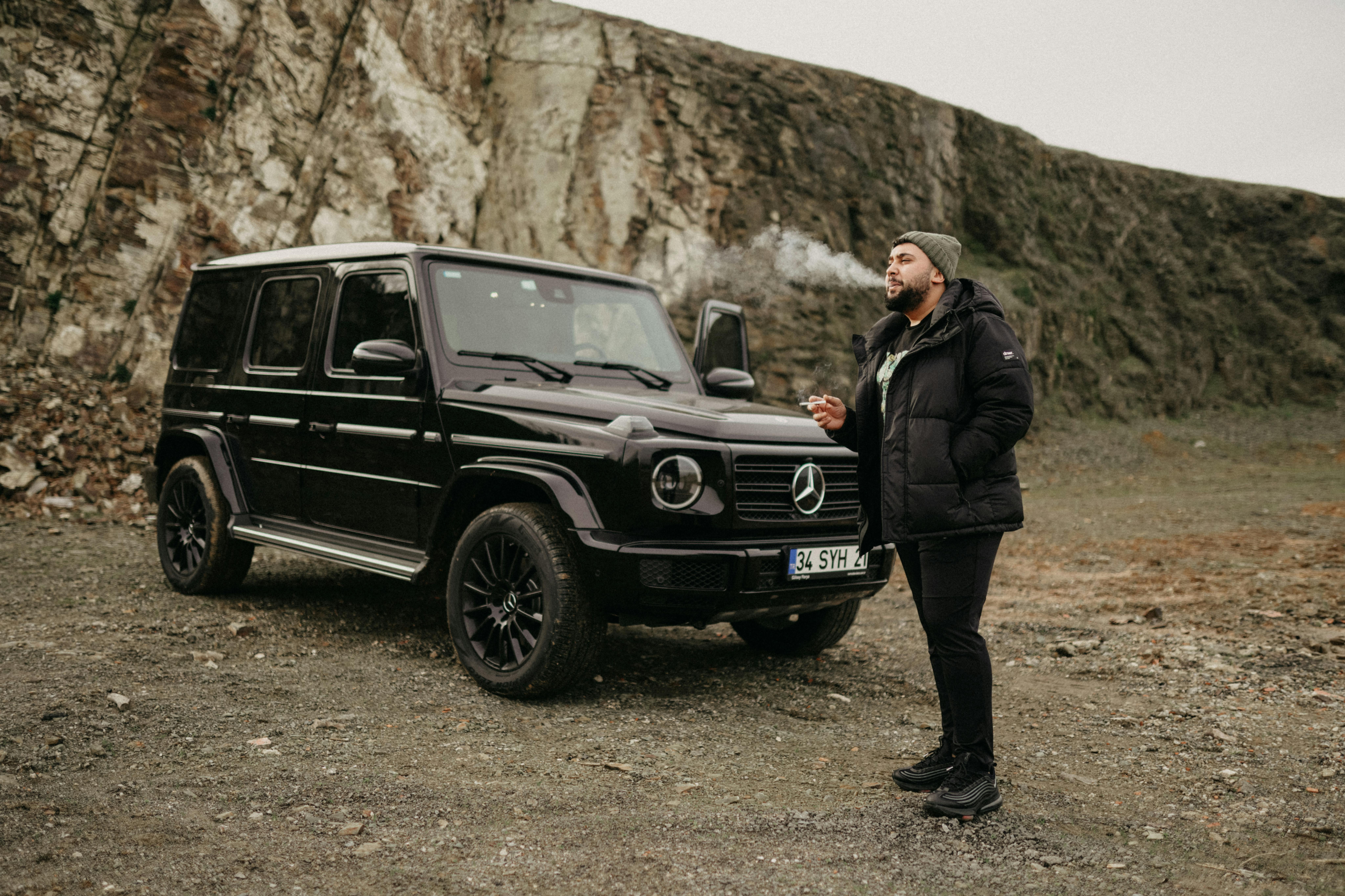 A man stands confidently next to a black SUV, set against rugged, rocky terrain.